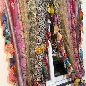Può includere: Una tenda colorata realizzata con scampoli di tessuto riciclati. La tenda presenta una varietà di motivi e colori, tra cui rosa, rosso, verde e nero. La tenda è appesa davanti a una finestra, aggiungendo un tocco bohémien alla stanza.