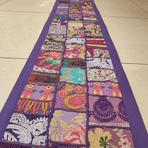 May include: A long, rectangular patchwork textile runner with a deep purple border. The center features a grid of colorful, embroidered squares in shades of purple, pink, gold, and teal. The runner is likely used for home decor.