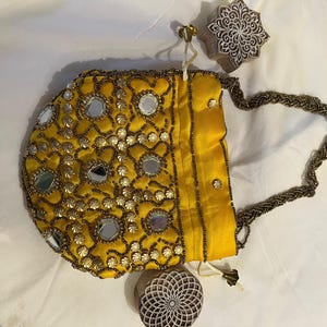 Op de afbeelding: Een gele, met kralen bezette handtas met kleine spiegels en een gevlochten handvat. De tas is versierd met goudkleurige kralen en pailletten. Twee houten stempels zijn ook zichtbaar.