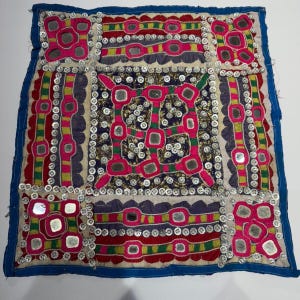 Può includere: Tessuto quadrato ricamato con bordo blu. Il design presenta un quadrato centrale blu scuro con motivi geometrici rosa, rossi e dorati, circondato da bianco e impreziosito da piccoli specchi e paillettes argentate. I bordi sono rifiniti con tessuto blu.