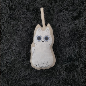 Può includere: Un giocattolo di peluche di gatto bianco con occhi blu e un semplice viso ricamato. Il gatto è appeso a un anello di corda.