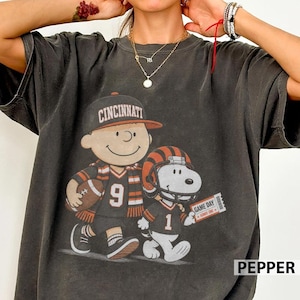 Könnte beinhalten: Ein anthrazitfarbenes Kurzarm-T-Shirt mit einer Grafik einer Zeichentrickfigur, die eine Cincinnati-Baseballkappe und ein Trikot mit der Nummer 9 trägt und einen Football hält, neben einem Hund mit Footballhelm und Trikot mit der Nummer 1.