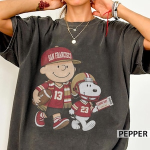 Könnte beinhalten: Ein anthrazitfarbenes Kurzarm-T-Shirt mit einer Grafik eines Zeichentrickcharakters, der eine San Francisco Baseballkappe und ein Trikot mit der Nummer 13 trägt, sowie einem Zeichentrickhund mit Footballhelm und Trikot mit der Nummer 23.