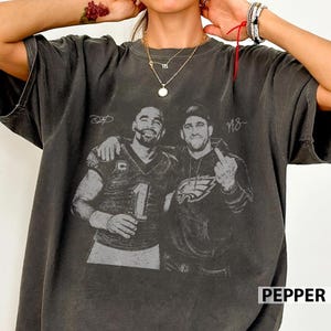 Op de afbeelding: Een antracietgrijs T-shirt met korte mouwen en een vintage zwart-wit grafische print van twee footballspelers. De ene speler draagt een shirt met het nummer 1, terwijl de andere een hoodie met een adelaarslogo draagt.