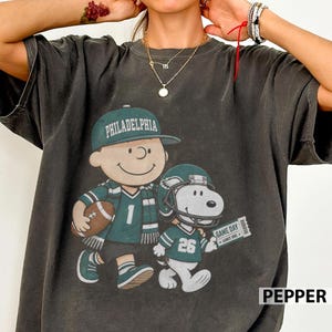 May include: A charcoal grey short-sleeve t-shirt featuring a graphic of a cartoon character wearing a Philadelphia cap and a football jersey with the number 1, alongside a cartoon dog wearing a football helmet and jersey with the number 26.