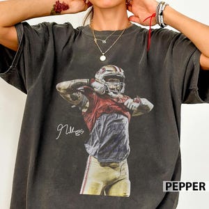 Może przedstawiać: Ciemnoszary t-shirt z krótkim rękawem z nadrukiem przedstawiającym zawodnika futbolu amerykańskiego w czerwonej koszulce, białym kasku i złotych spodniach. Zawodnik podnosi koszulkę, a po lewej stronie postaci widnieje biały podpis.
