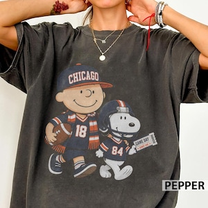 Könnte beinhalten: Ein anthrazitfarbenes T-Shirt mit einem Grafikdruck einer Zeichentrickfigur, die eine Chicago-Baseballkappe und ein Football-Trikot mit der Nummer 18 trägt, neben einem Zeichentrickhund mit Football-Helm und einem Trikot mit der Nummer 84.