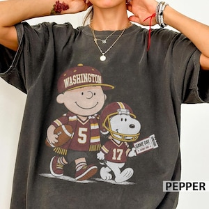 Pode incluir: Uma t-shirt cinzento-carvão de manga curta com um gráfico de uma personagem de desenho animado com um boné Washington e uma camisola com o número 5, segurando uma bola de futebol americano, ao lado de um cão com capacete e camisola 17.