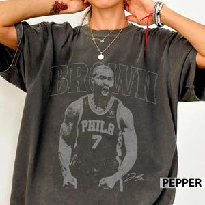 Pode incluir: Uma t-shirt de manga curta cinzento-carvão com uma grande impressão gráfica de um jogador de basquetebol com a camisola de Filadélfia e o número 7. A palavra BROWN está impressa em letras maiúsculas grandes acima do jogador e uma assinatura.
