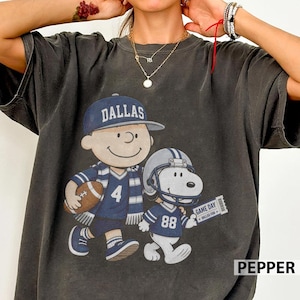 May include: A charcoal gray short-sleeve t-shirt featuring a graphic of a cartoon character wearing a blue Dallas cap and jersey with the number 4, holding a football, alongside a cartoon dog wearing a blue football helmet and jersey with the number 88.