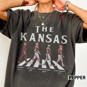 Op de afbeelding: Een antracietgrijs T-shirt met korte mouwen en de tekst THE KANSAS in grote witte letters boven een afbeelding van vier footballspelers die op een zebrapad lopen. De namen Andy Reid, Travis Kelce, Kenneth Walker III en Patrick Mahomes staan eronder.