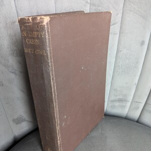 May include: A brown hardcover book titled The Men of the Last Frontier and An Empty Cabin by Grey Owl. The spine features gold lettering and shows signs of wear along the edges. The book is positioned vertically, displaying its front cover and spine.