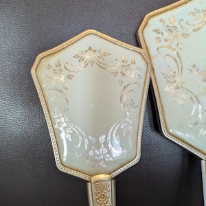 May include: Two vintage hand mirrors. The mirrors have a light-coloured, octagonal face with a floral design in gold and silver. The handles are silver with a decorative pattern.