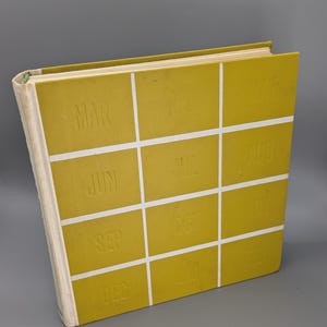 Op de afbeelding: Een geel boek met een rasterpatroon op de omslag. Elk vierkant heeft een maand van het jaar erop gedrukt, beginnend met maart en eindigend met februari.
