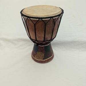 May include: A wooden djembe drum with a natural skin drumhead. The drum's body is a rich brown color, with a black rope net and a base decorated with colorful dots. The drum is designed for hand percussion.