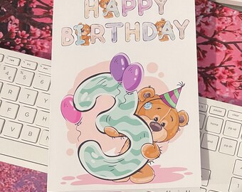 May include: A birthday card with the words "HAPPY BIRTHDAY" in pastel colors. A cartoon teddy bear wearing a party hat hugs a large number "3" with purple balloons. The card is on a desk with a keyboard.