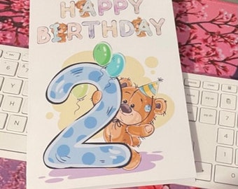 May include: A birthday card with the words "HAPPY BIRTHDAY" at the top, decorated with teddy bears. A large blue number "2" with white polka dots is in the center, with a cartoon teddy bear hugging it. Green and blue balloons are above the bear.