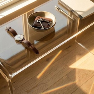 Puede incluir: Una mesa auxiliar de acrílico transparente con bordes redondeados. Un reloj de cuero marrón con esfera plateada descansa sobre la mesa. Un cuenco pequeño contiene una cartera y llaves. Un bolígrafo y un bloc de notas también están sobre la mesa.