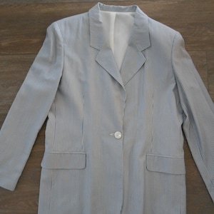 Può includere: Una giacca blu chiaro a righe bianche. La giacca ha una chiusura con un solo bottone e due tasche.