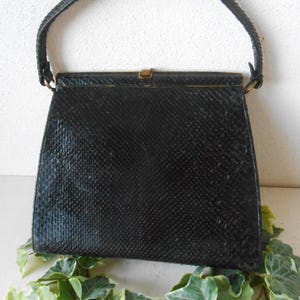 Könnte beinhalten: Eine schwarze Vintage-Handtasche mit strukturiertem Muster und gebogenem Griff. Die Tasche hat eine rechteckige Form mit einem Verschluss und goldfarbener Hardware. Der Griff ist ebenfalls schwarz und scheint aus einem ähnlichen Material wie die Tasche zu bestehen.