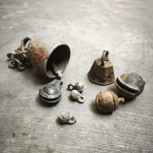 Op de afbeelding: Een verzameling antieke metalen bellen met roest- en slijtagesporen. Een grote bel met ketting ligt op zijn kant, omringd door kleinere bellen in verschillende vormen. Op een grijze ondergrond.