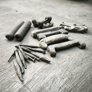 Może przedstawiać: Asortyment antycznych metalowych okuć, w tym gwoździe, uchwyty i elementy cylindryczne, ułożone na teksturowanej szarej powierzchni. Przedmioty mają wygląd zwietrzały, sugerujący historyczne użytkowanie i rzemiosło.