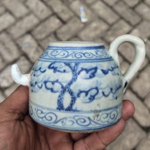 Può includere: Piccola teiera antica in ceramica con base bianca e motivi floreali blu. La teiera ha un manico curvo e un beccuccio. Il design include un motivo ad albero e nuvole. La teiera è tenuta in mano.
