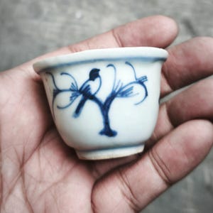 Op de afbeelding: Kleine witte keramische theekop met een blauw vogel- en takontwerp. De kop wordt in een hand gehouden, wat het delicate kunstwerk laat zien. Het blauwe ontwerp contrasteert met het witte keramiek en creëert een klassieke esthetiek.