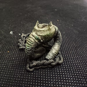 Puede incluir: Una escultura de metal verde y marrón de una criatura con una cadena envuelta alrededor. La escultura está sobre una superficie negra texturizada.