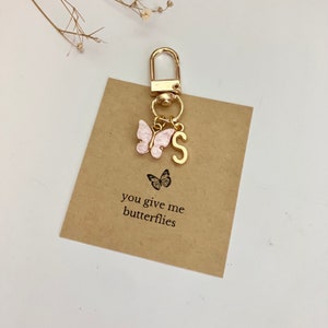 May include: Gold keychain with a pink butterfly charm and a gold letter 'S' charm. The keychain is attached to a brown card with the text 'you give me butterflies'.