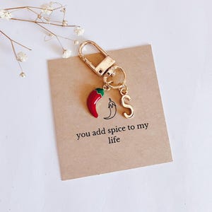 May include: A gold keychain with a red chili pepper charm and a gold letter "S" charm. The keychain has a tag that reads "you add spice to my life".