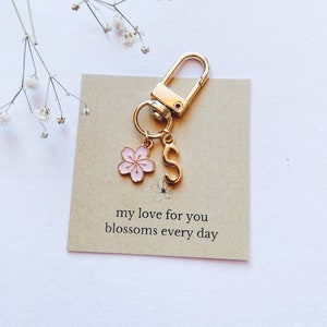 May include: Gold keyring with a pink flower charm and a gold letter 'S' charm. The keyring is on a brown card with the text 'my love for you blossoms every day'.