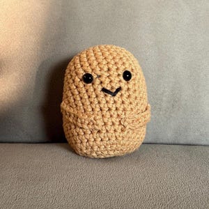 Puede incluir: Un juguete de peluche de patata tejido a crochet con una cara sonriente y ojos negros. La patata es marrón y tiene una superficie texturizada.