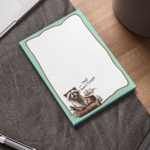 Op de afbeelding: Een rechthoekig notitieblok met een turquoise rand en een wasbeerillustratie. Het notitieblok heeft de tekst "- Hey! Don't Forget" en ligt op een donkergrijze ondergrond.