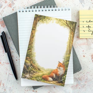 May include: A notepad with a forest scene and a fox on the cover, alongside a black pen and a grey notebook. The notepad has lined pages and a spiral binding. A yellow sticky note with handwritten text is also visible.