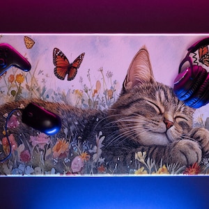 Peut inclure: Tapis de souris rectangulaire avec un chat tigré endormi dans un champ de fleurs. Une manette de jeu, une souris et un casque noirs sont également représentés. Des papillons voltigent autour du chat. Le tapis de souris a un bord lumineux.