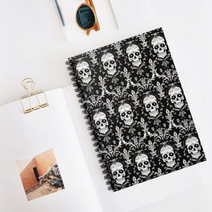 May include: A spiral-bound notebook with a black cover featuring a repeating pattern of white skulls and floral designs. The notebook is open on a white surface, with a gold paper clip and sunglasses visible.