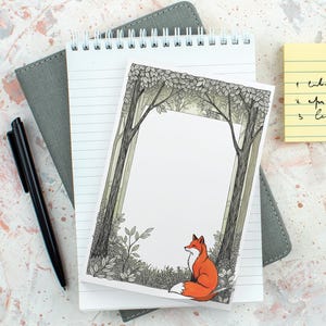 May include: A notepad with a forest scene illustration and a red fox, alongside a black pen and a grey notebook. The notepad has lined paper and a spiral binding. A yellow sticky note is in the background.