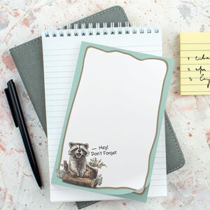 May include: A notepad with a raccoon illustration and the text "Hey! Don't Forget". The notepad is on top of a lined notepad and a gray notebook. A black pen is also present.