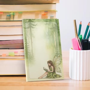 May include: A notepad featuring a forest scene and a fairy reading. The notepad has a green border and a watercolour illustration. Pencils, pens, and an eraser are in a white ceramic holder next to a stack of books on a wooden surface.