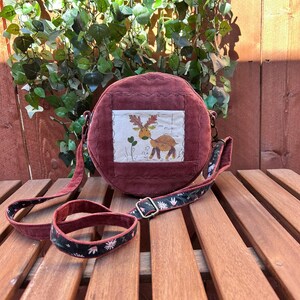 May include: A burgundy corduroy crossbody bag with a stitched deer design on the front. The bag has a matching burgundy strap and a patterned strap with floral accents. The bag is round and sits on a wooden surface.