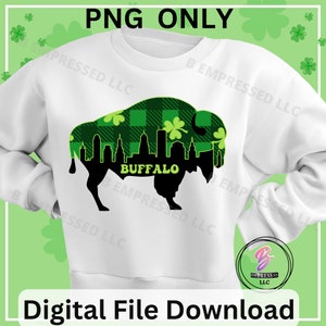 May include: A white sweatshirt with a black buffalo silhouette design. The buffalo is filled with a green and black plaid pattern and green shamrocks. The text "BUFFALO" is written in black below the buffalo. The text "PRESSED LLC" is written in black on the sleeves and bottom of the sweatshirt.