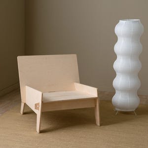 May include: A light wood chair with a simple design sits on a beige rug in front of a white paper lamp.