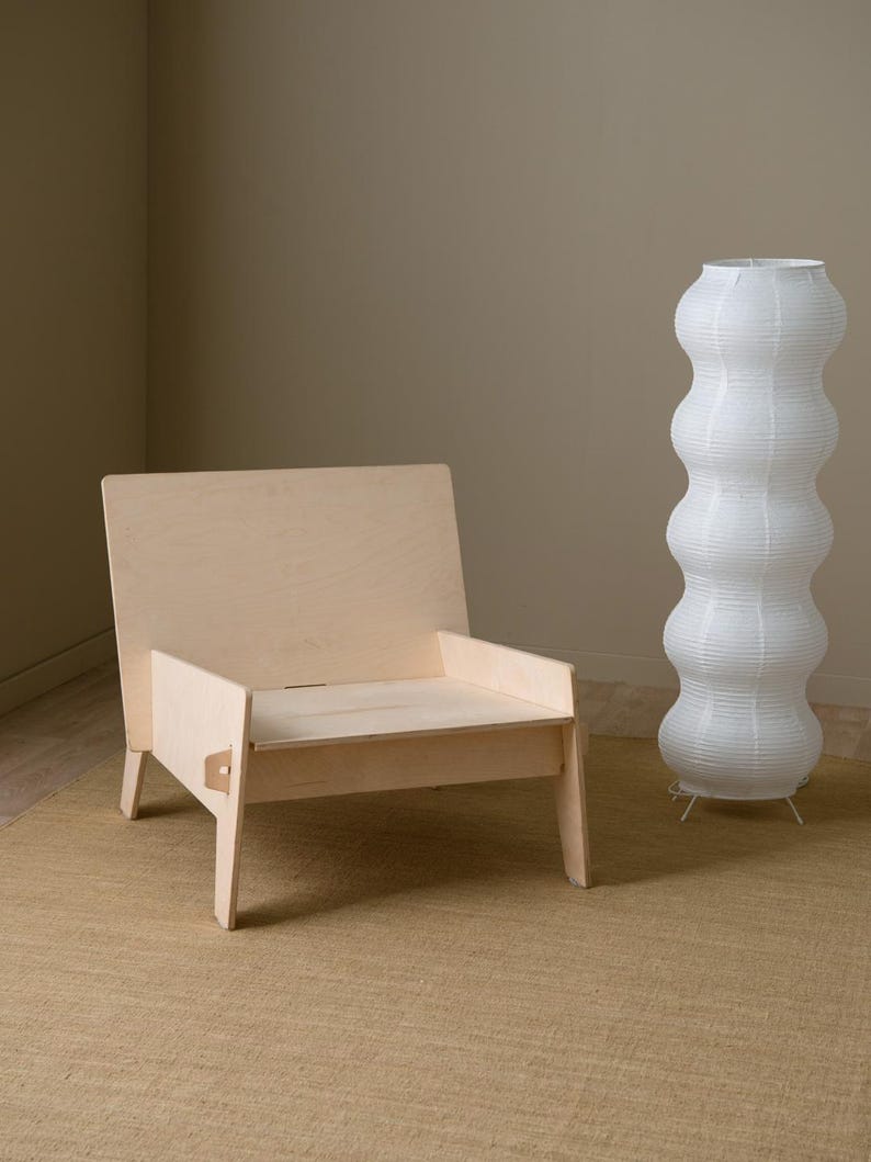 May include: A light wood chair with a simple design sits on a beige rug in front of a white paper lamp.