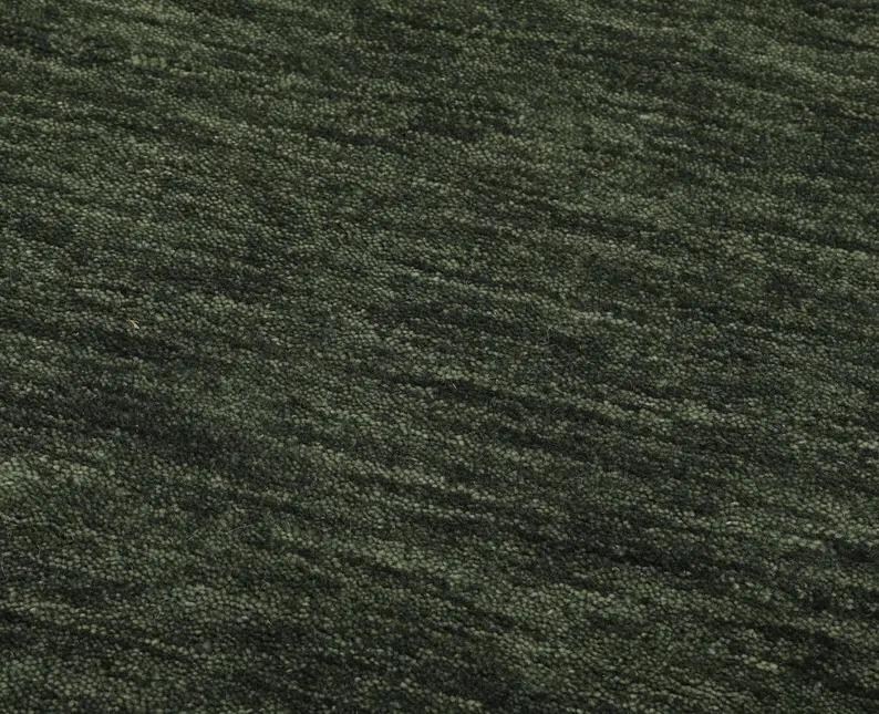 May include: Close-up of a textured, dark green rug. The rug has a dense, looped pile with a subtle, diagonal pattern. The color is a deep, muted green, with variations in tone creating visual interest. The rug appears soft and inviting.