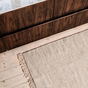 May include: A light brown rug with fringed edges, laid out on a wooden floor. The rug is in front of a dark brown wooden wall.