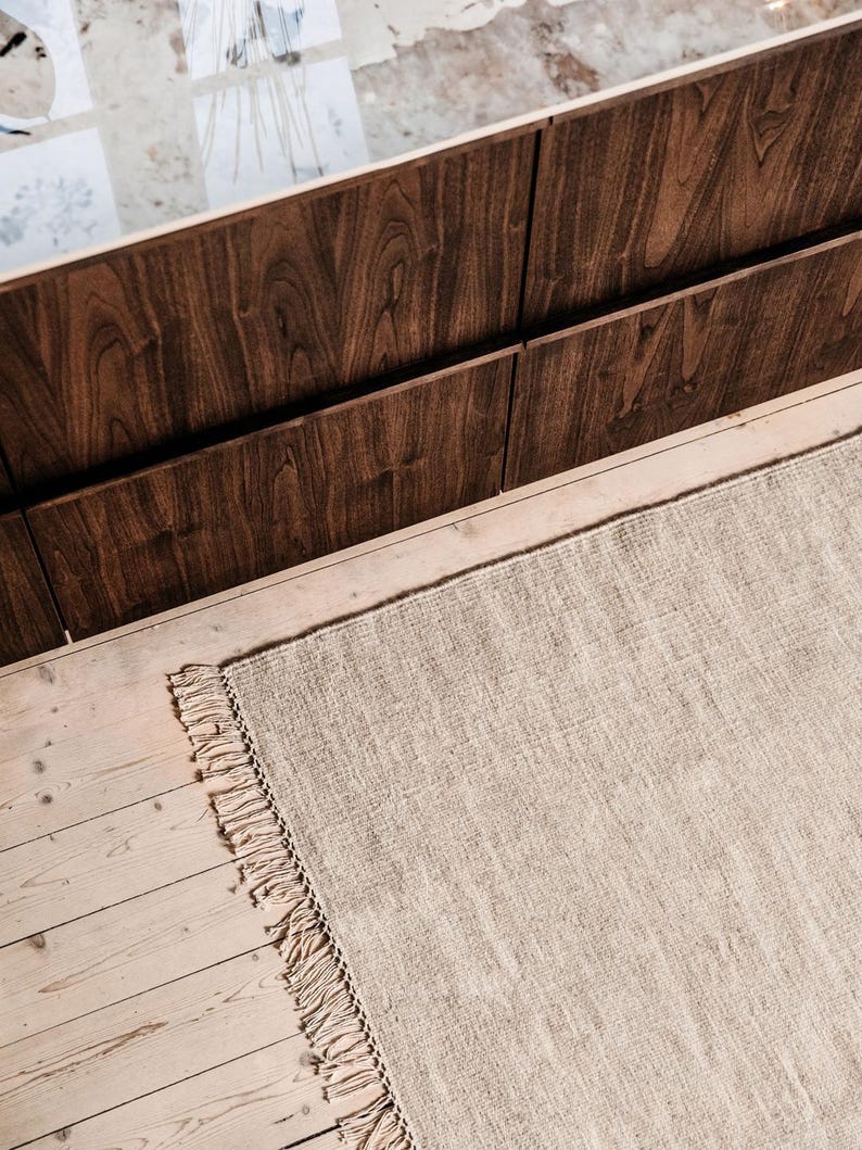 May include: A light brown rug with fringed edges, laid out on a wooden floor. The rug is in front of a dark brown wooden wall.