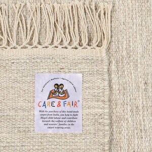 May include: A beige hand-woven rug with a fringe border. The rug has a label that reads "CARE & FAIR" and explains that the purchase of the rug helps fight illegal child labour in India.
