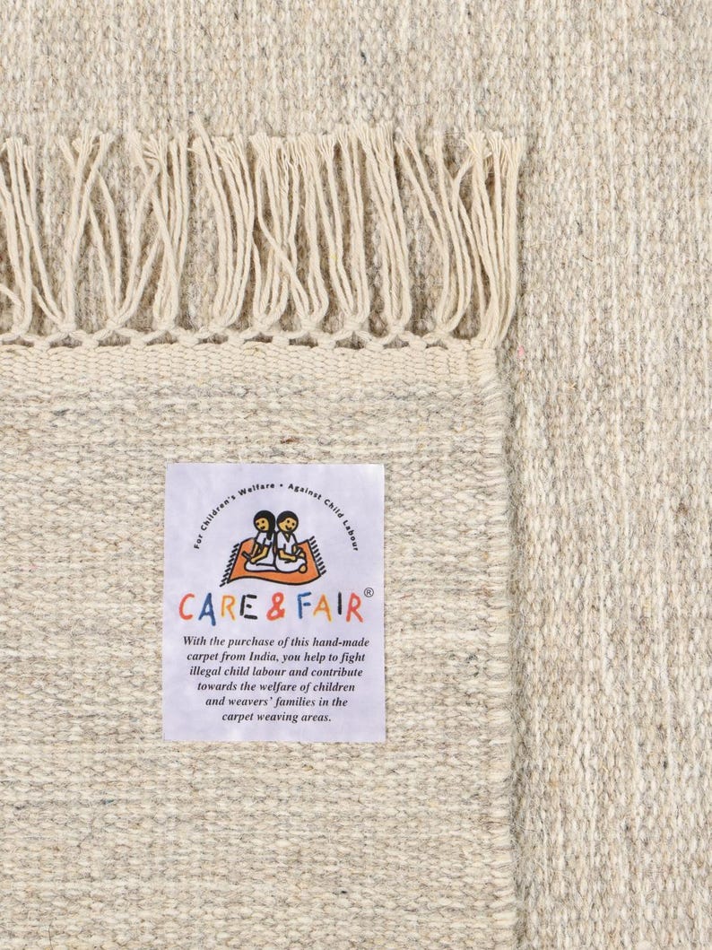 May include: A beige hand-woven rug with a fringe border. The rug has a label that reads "CARE & FAIR" and explains that the purchase of the rug helps fight illegal child labour in India.