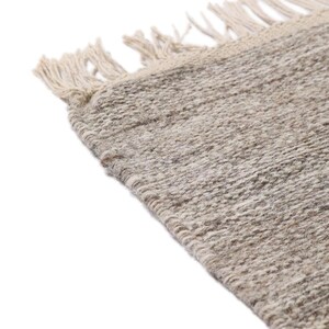 May include: Close-up of a beige woven rug with fringe on the edges. The rug has a subtle texture and a natural, earthy tone.